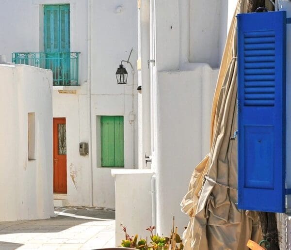 In the quaint village of Lefkes, Paros, Greece, a narrow street lined with white buildings and blue shutters is serenely brightened by a sunlit path where a small black cat gracefully strolls.