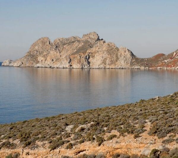 A rocky coastline on Tilos, Greece, features calm blue waters and a hilly landscape. In the foreground, a weathered stone structure adds rustic charm to this scenic vista.