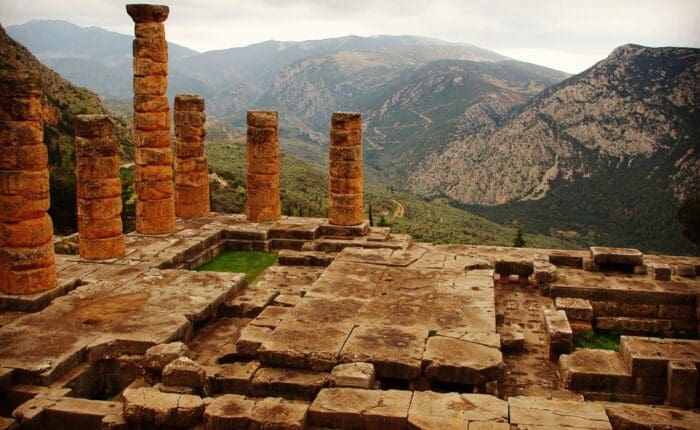 Ancient stone ruins and columns grace a mountainous landscape, evoking the mystique of once-vibrant royal sites. The hills and cloudy sky in the background whisper secrets of past oracles and gods, leaving an air of timeless wonder.