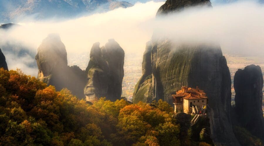 Perched like an oracle on a rocky pillar, the monastery overlooks a sea of mist and vibrant fall foliage, with high cliffs forming a majestic backdrop.