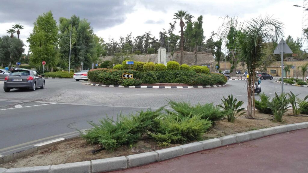 The circular roundabout in Nicosia, Cyprus, is bustling with cars amidst lush greenery and palm trees. Set against an overcast sky, visible traffic signs guide your journey. A visit here is a must on any travel guide of things to do in Nicosia.