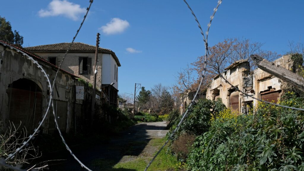 A barbed wire fence frames an abandoned village street in Nicosia, Cyprus, with dilapidated buildings and overgrown vegetation under a bright blue sky—a haunting yet captivating scene for any travel guide.