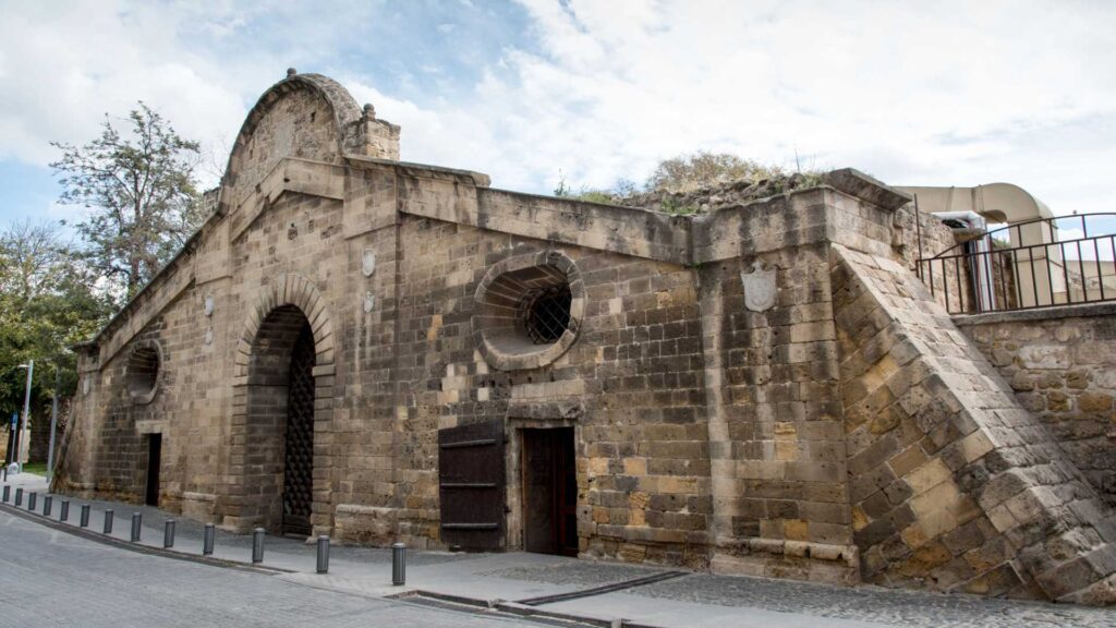 Nestled on a cobblestone street in Nicosia, this historic stone building boasts arched entrances and barred windows. Stone crest emblems adorn the walls, set against a backdrop of Cyprus' trees and sky, as if lifted from a travel guide's pages.