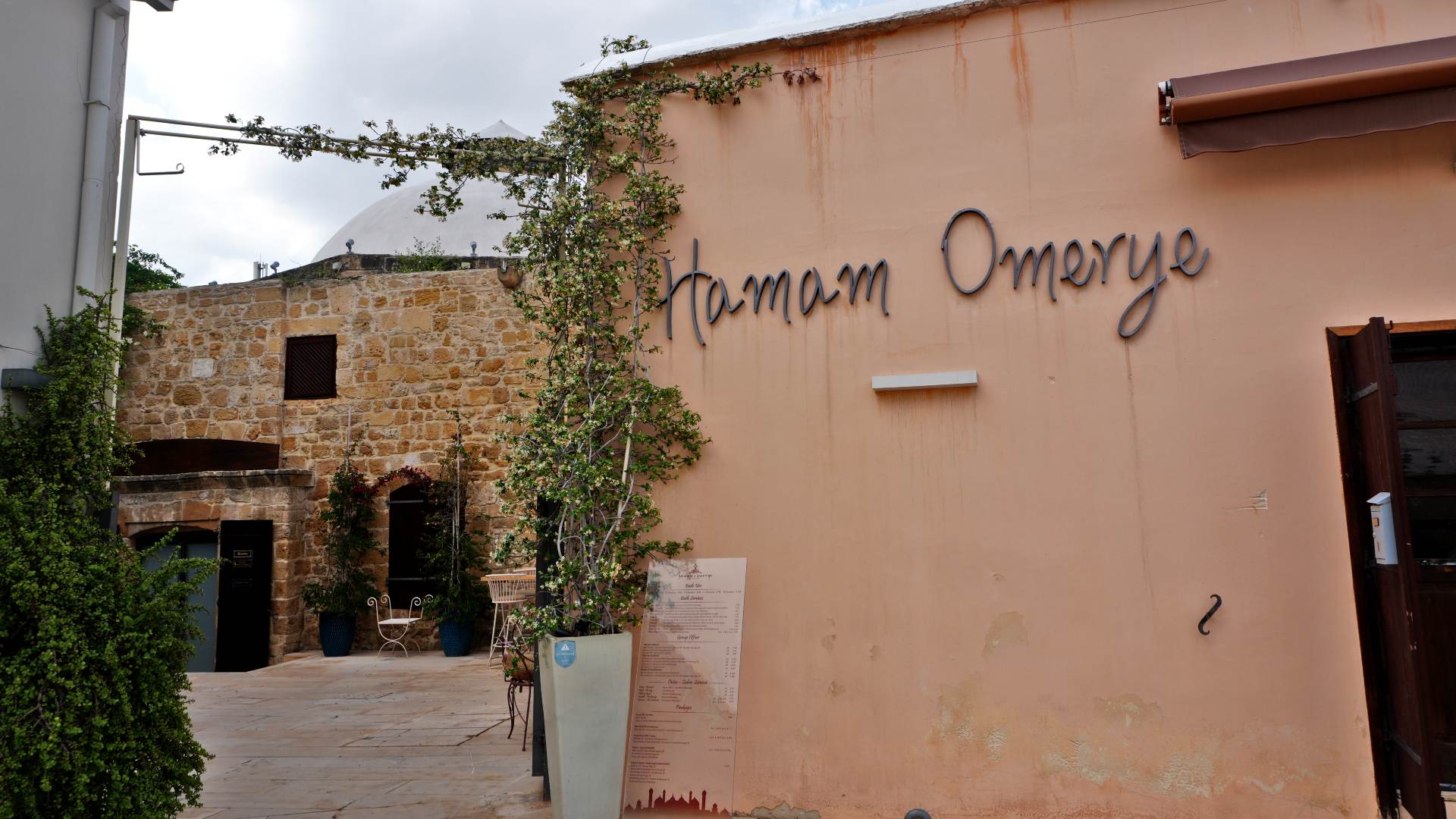 Exterior view of Hamam Omerye, a must-see when exploring what things to do in Nicosia Cyprus—featuring a historic stone building, peach-colored wall with its name, lush greenery, and an inviting entrance with a signboard - Hamam Omerye, Nicosia, Cyprus