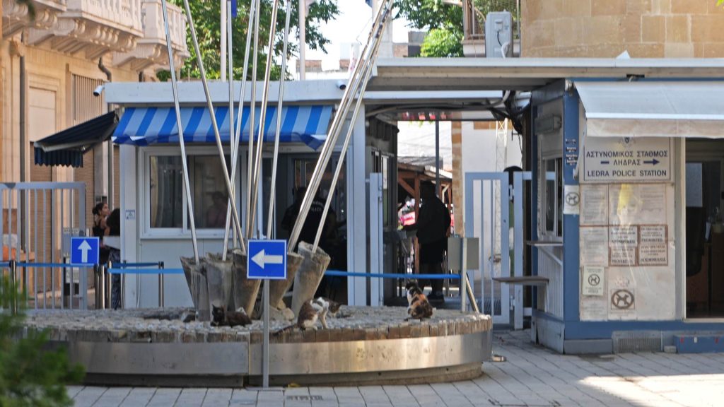 Pedestrian border checkpoint with flagpoles, directional signs, and a police station kiosk—an essential crossing for visitors exploring what things to do in Nicosia, Cyprus as people pass through the entrance.