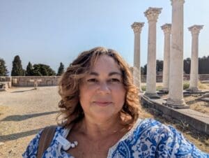 A person with sunglasses on their head stands outdoors in front of ancient stone columns, wearing a blue and white patterned shirt.