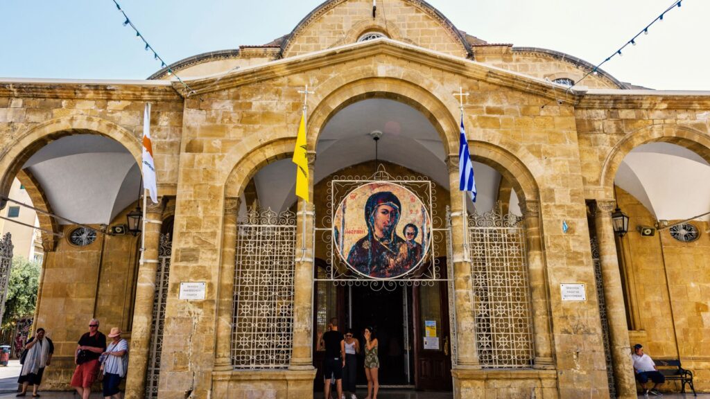 Discover the majestic stone church entrance in Nicosia, Cyprus, featuring arched doorways and a revered religious icon. Colorful flags sway as visitors gather, with some venturing inside. Perfectly capturing the essence of local culture, it's a must-see on any travel guide itinerary.