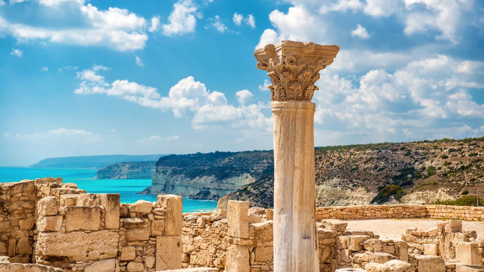 Ancient stone column amidst ruins with a backdrop of cliffs and turquoise sea under a blue sky with clouds.