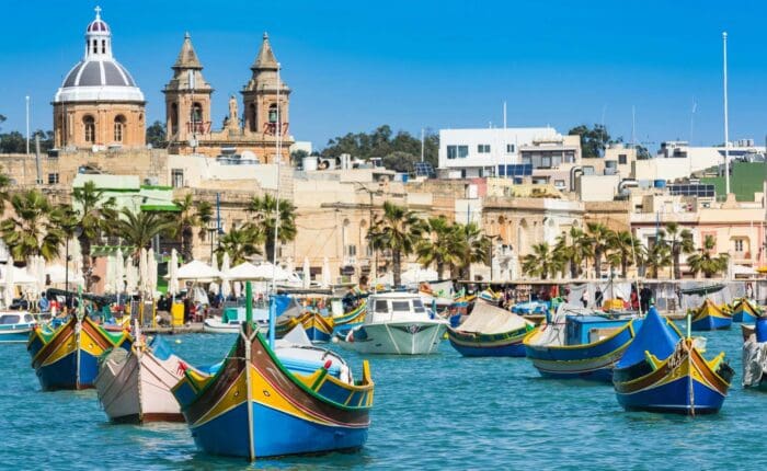 Colorful traditional boats float in a harbor with historic buildings and churches in the background under a clear blue sky—a picturesque scene often enjoyed on Cyprus tours.