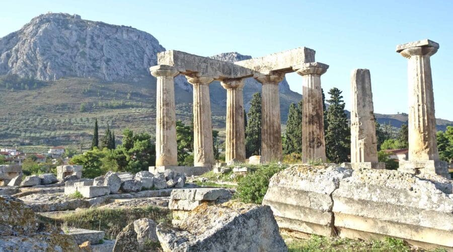 Ruins of ancient stone columns and structures stand in front of a mountain, surrounded by scattered stones and greenery under a clear sky—a perfect scene to explore on a Peloponnese tour.