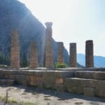 Ancient stone columns and ruins of the Temple of Apollo at Delphi, Greece, stand against a backdrop of mountains and trees—a must-see on any tour calendar.