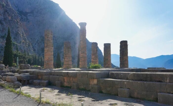Ancient stone columns and ruins of the Temple of Apollo at Delphi, Greece, stand against a backdrop of mountains and trees—a must-see on any tour calendar.