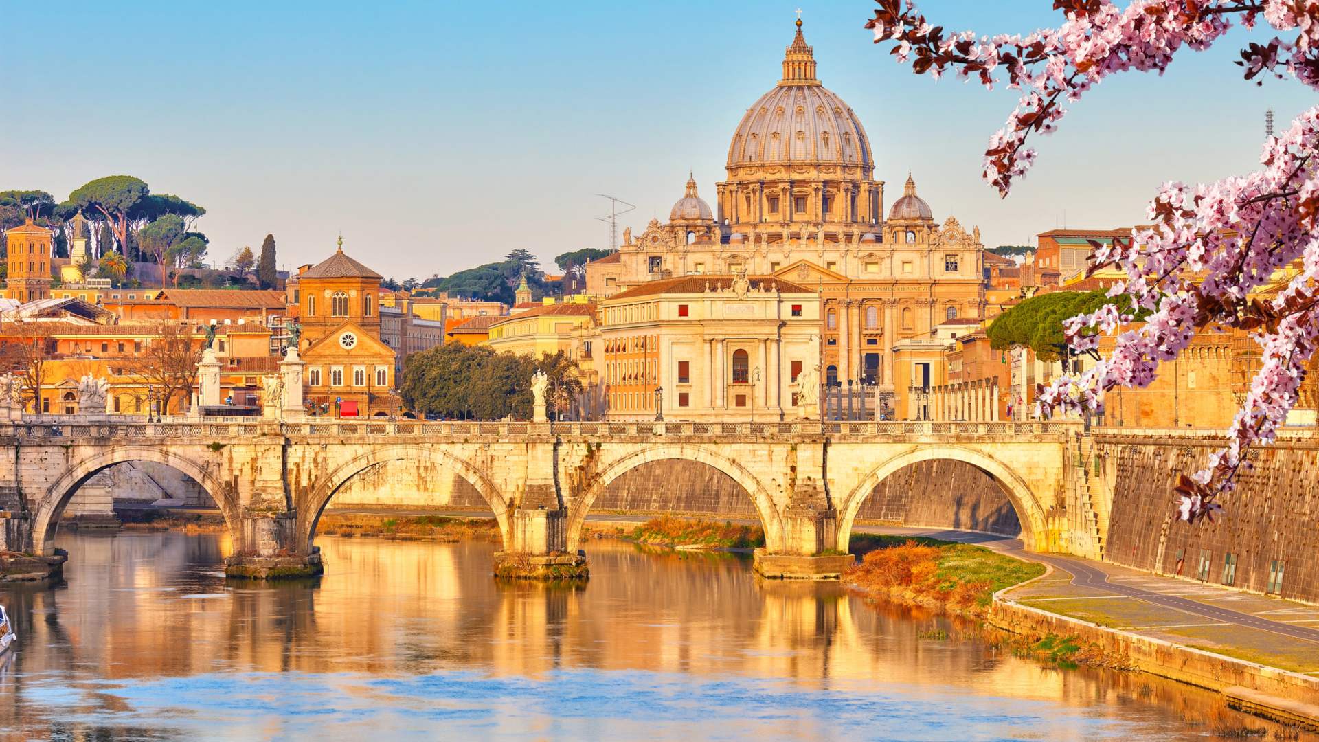 Enjoy a stunning view of St. Peter’s Basilica and the Vatican from across the Tiber River in Rome, framed by a stone bridge and blooming cherry blossoms—perfect inspiration for custom trips to Italy.