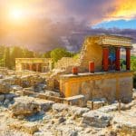 Explore the ancient ruins of the Knossos Palace in Crete, Greece, with stone walls, red columns, and trees beneath a dramatic sunset sky—a must-see highlight on any Greece tour.