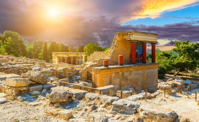 Explore the ancient ruins of the Knossos Palace in Crete, Greece, with stone walls, red columns, and trees beneath a dramatic sunset sky—a must-see highlight on any Greece tour.