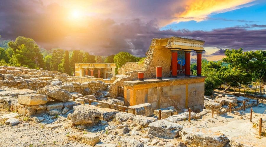Explore the ancient ruins of the Knossos Palace in Crete, Greece, with stone walls, red columns, and trees beneath a dramatic sunset sky—a must-see highlight on any Greece tour.