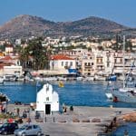 Aegina Island, Greece - Charming small white chapel on a waterfront pier in Aegina Island, with people strolling nearby, sailboats docked in the marina, and a hillside town beyond—a perfect Athens island day trip spot for your 2026 guide.