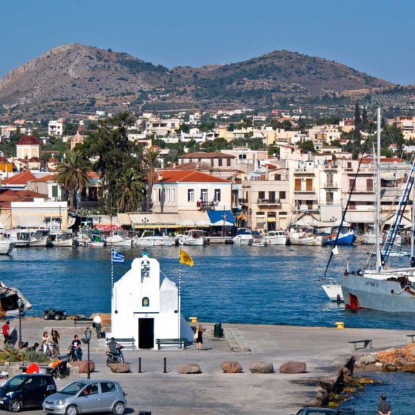 Aegina Island, Greece - Charming small white chapel on a waterfront pier in Aegina Island, with people strolling nearby, sailboats docked in the marina, and a hillside town beyond—a perfect Athens island day trip spot for your 2026 guide.