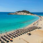 Aerial view of a sandy beach with sun loungers and umbrellas, turquoise water, people swimming, and hidden coves nearby—typical of Peloponnese beaches—with a small island in the background under a clear blue sky.