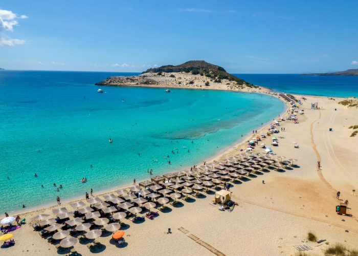 Aerial view of a sandy beach with sun loungers and umbrellas, turquoise water, people swimming, and hidden coves nearby—typical of Peloponnese beaches—with a small island in the background under a clear blue sky.