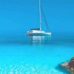 A white sailboat floats on calm, clear turquoise water near rocky shorelines under a bright blue sky, offering a glimpse of the serene beauty you can experience on Paros Boat Tours.