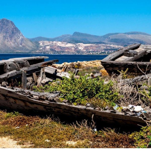 Two old, decaying wooden boats rest on dry land with grass growing inside, overlooking the sea and distant mountains under a clear blue sky—an evocative scene on a Sicily tour.
