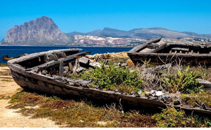 Two old, decaying wooden boats rest on dry land with grass growing inside, overlooking the sea and distant mountains under a clear blue sky—an evocative scene on a Sicily tour.