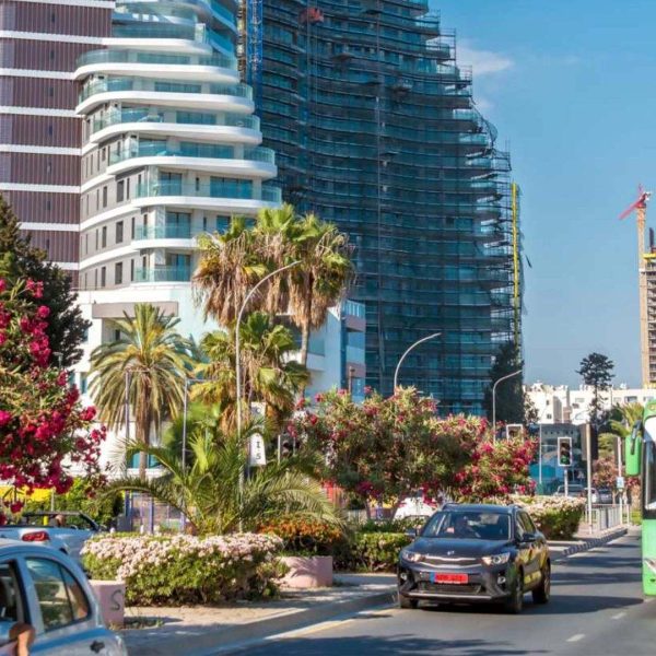 A city street with cars and a green intercity coach, possibly a Paphos to Limassol coach, lined with flowering trees and modern high-rise buildings in the background.