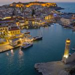 Aerial view of the harbour in Rethymno, Crete, with boats gently bobbing along the waterfront, surrounding buildings lit at dusk, a lighthouse standing proudly on a stone pier, and a hilltop fort commanding the background.