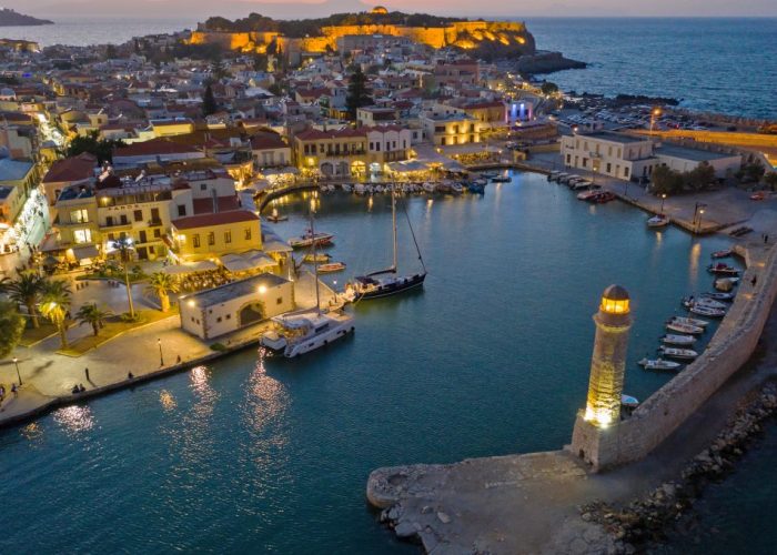 Aerial view of the harbour in Rethymno, Crete, with boats gently bobbing along the waterfront, surrounding buildings lit at dusk, a lighthouse standing proudly on a stone pier, and a hilltop fort commanding the background.